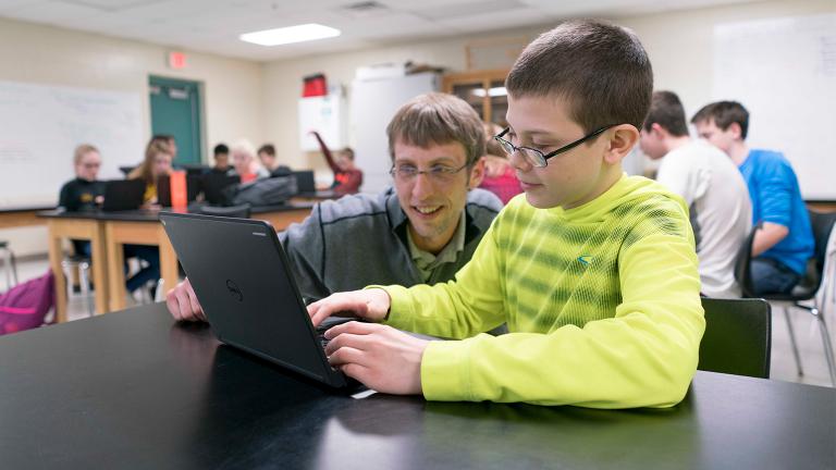 teacher crouched at a student's desk assisting with a project on a laptop